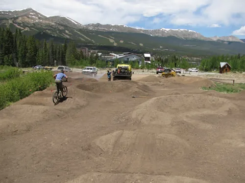 Mountain biker riding the rollers of a newly built dirt pump track, with snow-streaked peaks behind and a yellow tracked machine working at the top edge of the site