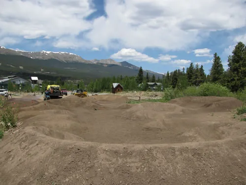 Freshly shaped dirt pump track with rollers and berms, backed by a snow-streaked mountain range under tall cumulus clouds, with a yellow compact loader parked at the far edge