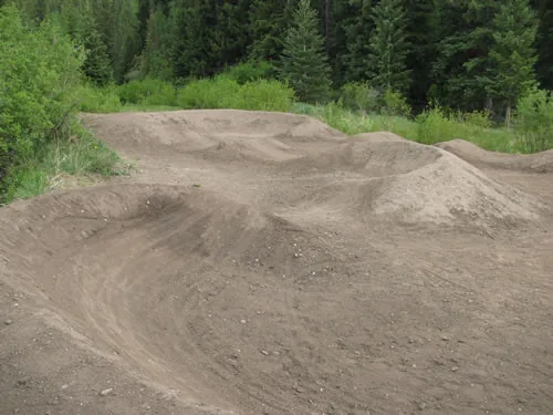 Sculpted dirt pump track berms and rollers with smoothly finished surfaces, backed by a dense green treeline