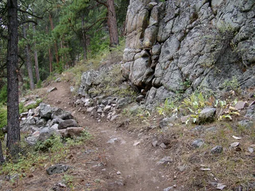 Narrow dirt singletrack running tight against a tall fractured granite outcrop, with a dry-stacked stone edge supporting the downhill side of the trail