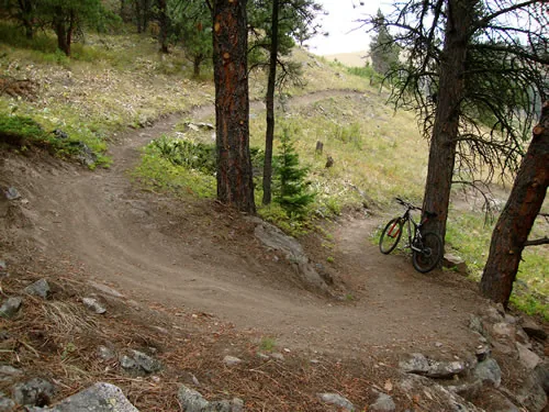 Wide bermed switchback turn on dirt singletrack in an open pine stand, with a mountain bike leaning against a tree beside the trail