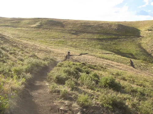 Singletrack climbing across a broad open grass and sagebrush hillside in low warm light, with a small distant figure on the trail ahead
