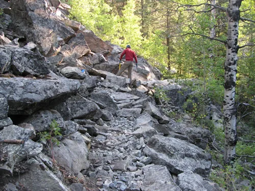 Trail worker in a red shirt holding a long rock bar while standing on a steep granite boulder field above a section of stone-armored trail