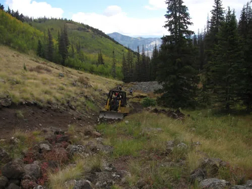 A small yellow tracked trail dozer cutting a new trail bench across a steep grassy slope above a forested valley