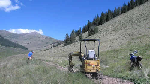 A yellow mini-excavator on a newly cut trail bench in an open alpine valley, with a worker using a hand tool nearby under a blue sky