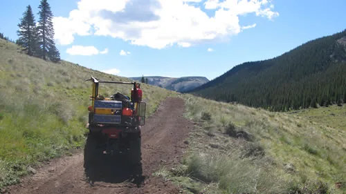 A red trail machine driving up a newly cut trail bench through an open alpine valley under a blue sky with scattered clouds