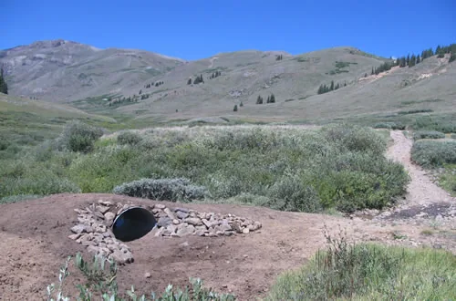 A culvert outlet with a hand-placed stone headwall built into a trail crossing in a high open basin, with a narrow trail visible beyond