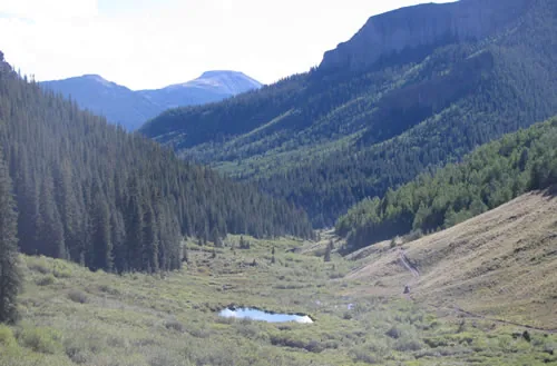 A deep mountain valley with layered cliff bands, dark conifer slopes and a small pond on the valley floor