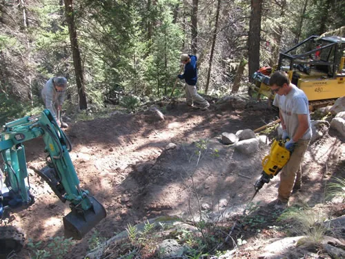 Trail crew working a rocky forest hillside with a mini excavator and a handheld pneumatic rock drill