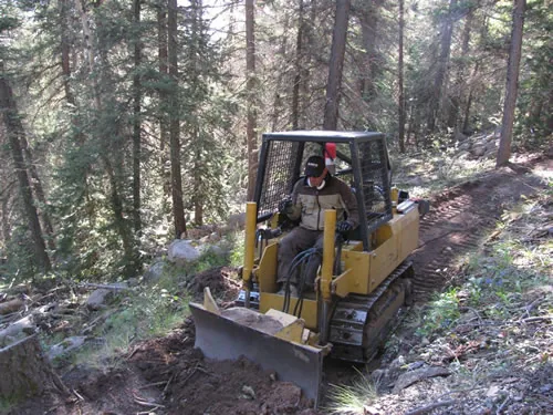 Small tracked trail dozer with a front blade cutting new bench tread across a steep forested hillside