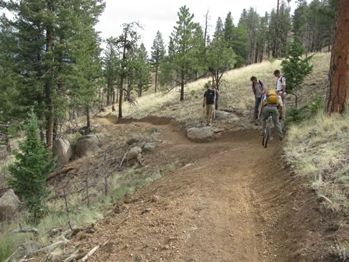 A wide, freshly finished singletrack trail sweeping up a grassy hillside with three people and a bike near the upper turn