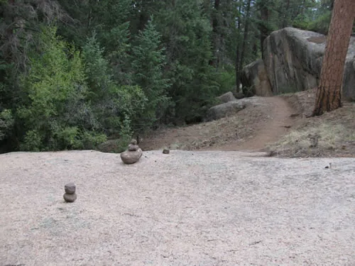 Trail crossing a broad granite slab marked with small stacked rock cairns before entering the conifer forest