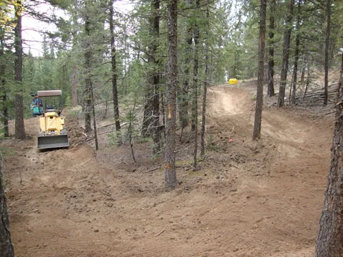 Small tracked trail dozer parked at the edge of a wide, freshly cut trail corridor in a lodgepole forest
