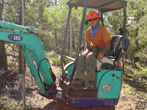 An operator in an orange hard hat runs a small teal mini excavator on a forest trail