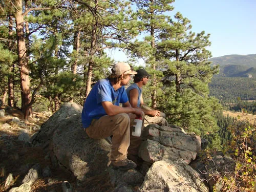 Two crew members sit on a granite outcrop in warm evening light looking out over a forested valley