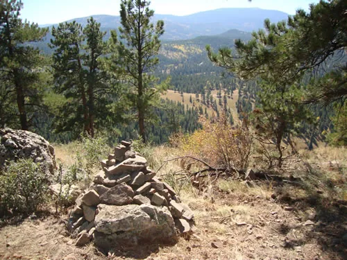 A stacked stone cairn stands on a rocky ridge above a forested valley with mountains beyond