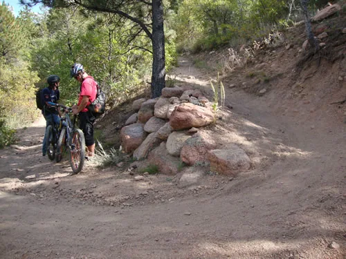 Two mountain bikers pause beside a pink granite retaining wall built along a dirt trail junction