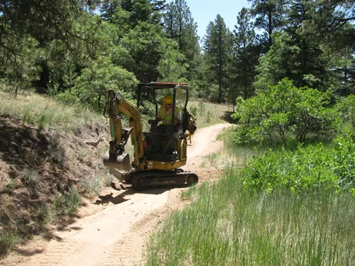 A mini excavator sits on a freshly cut dirt singletrack winding through a green pine forest