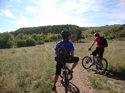 Two mountain bikers ride away from the camera on a singletrack trail through a grassy meadow toward rolling hills
