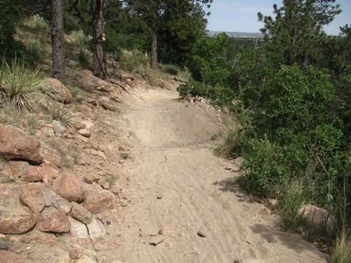 A dirt trail climbs beside a stacked pink granite retaining wall with yucca and scrub oak alongside