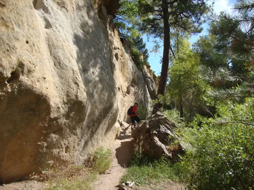 A narrow trail runs along the base of a tall pale sandstone cliff with a hiker in a red shirt beside it
