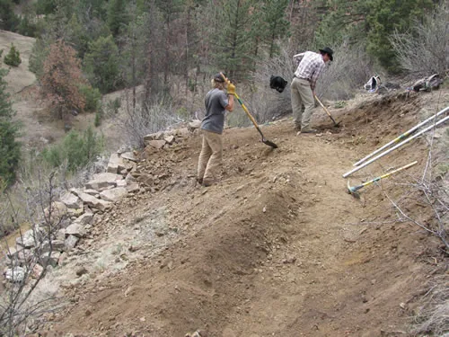 Two trail workers using hand tools on a fresh bench cut across a steep slope, with a dry-stacked stone retaining wall alongside