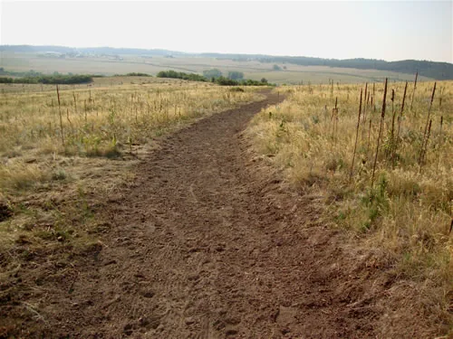 Freshly cut trail corridor of raw dirt running through dry prairie grass toward low hazy hills
