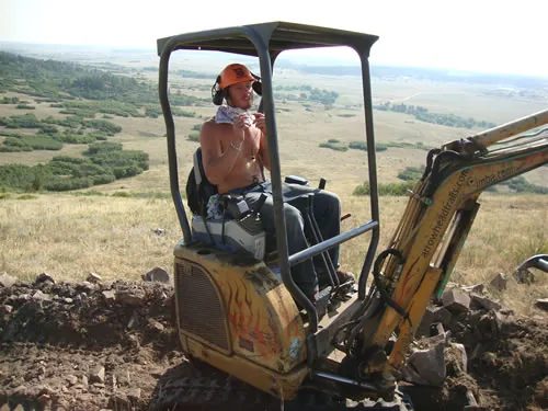 Operator in a hard hat running a small tracked mini-excavator cutting trail on a ridge above a broad valley