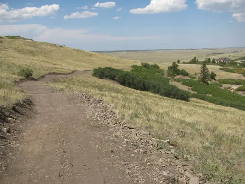 Bench-cut dirt trail traversing a grassy hillside with a rock-armored downhill edge above open plains