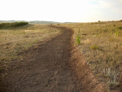 Freshly cut dirt trail curving through dry prairie grass in warm low evening light