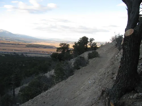 Trail cutting across a steep ridge shoulder at dusk above a wide valley, with a distant snow-dusted mountain range on the horizon