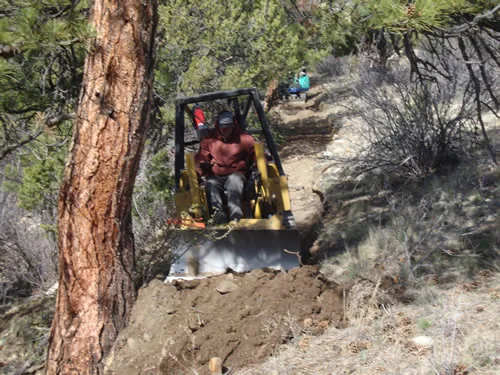 A yellow tracked trail dozer with an operator cuts a fresh singletrack bench across a steep pine-covered slope, with the new tread curving away uphill