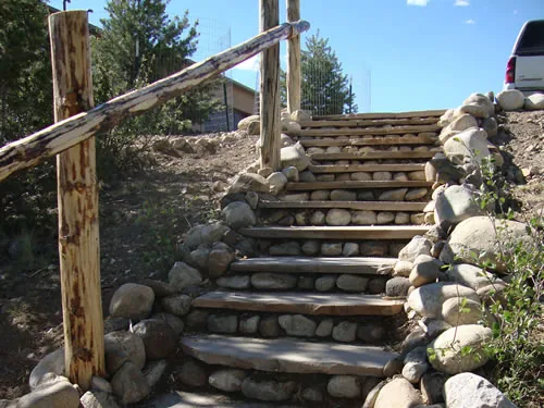 A stairway of timber treads set between dry-laid river cobble walls climbs a slope beside a peeled log handrail