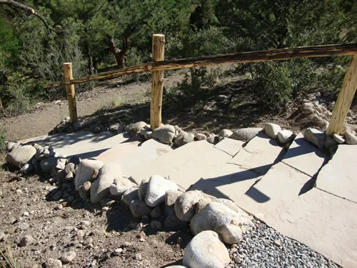 A wide smooth accessible path curves downhill edged with large placed cobbles and bordered by a peeled log post-and-rail fence among pinyon and juniper