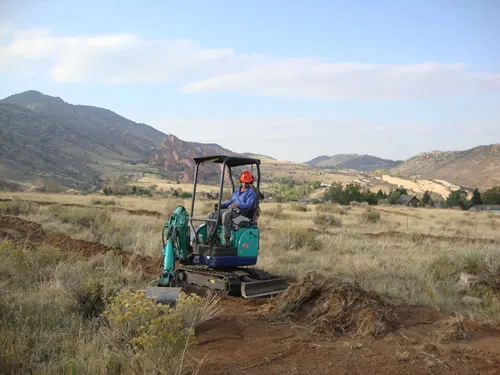 Operator in an orange hard hat running a small tracked excavator to cut a new trail bench across an open grassland foothill, with rocky ridges and red sandstone outcrops in the distance