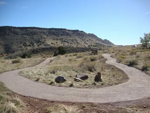 A tight switchback of finished crusher-fine trail loops around a cluster of placed boulders below a long mesa ridge