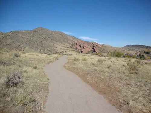 A crusher-fine trail curves through dry grassland toward red sandstone fins and a grassy foothill under a deep blue sky
