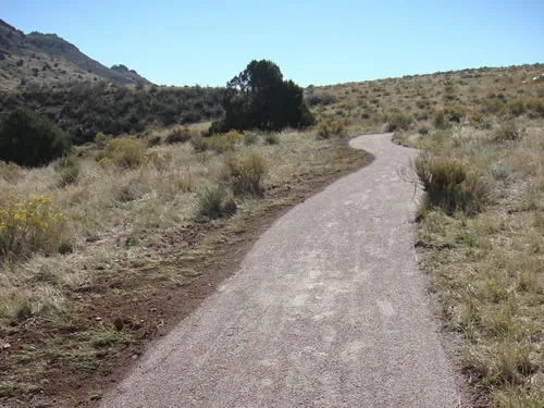 A crusher-fine trail rises and curves away between rabbitbrush and dry grass, with a juniper on the skyline and a scrubby hillside at left