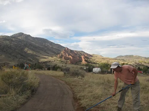 A worker in a ball cap drags a rake along a freshly built crusher-fine trail, with a grassy foothill, red rock outcrops and a broad cloudy sky behind
