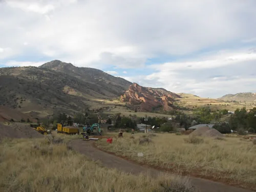 A trail runs through foreground grassland toward a valley with parked yellow trail equipment at left and red sandstone fins rising below a heavy cloudy sky