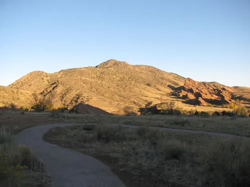 A sweeping S-curve of finished crusher-fine trail runs through dry grassland below a golden-lit ridge and red rock fins at sunset