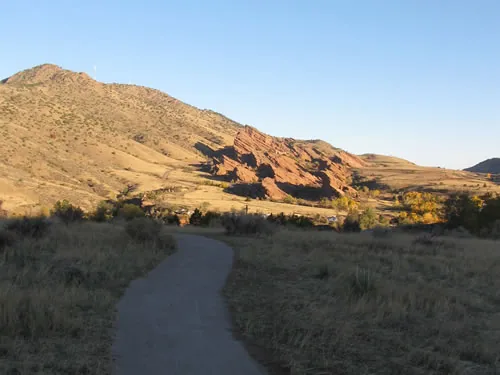 A smooth crusher-fine trail curves through golden autumn grassland toward tilted red sandstone fins and a sunlit ridge under a clear sky