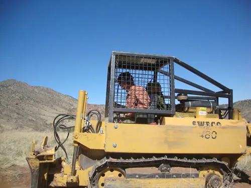A yellow tracked trail dozer with a caged rollover canopy sits on a dry hillside, its operator seated at the controls under a clear blue sky