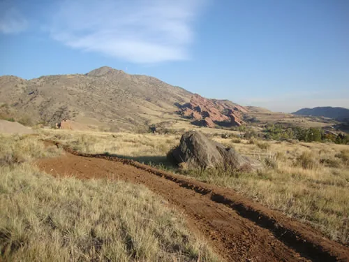 A freshly cut raw dirt trail corridor curves through dry grassland past a large boulder toward a pointed peak and red sandstone fins under wispy clouds