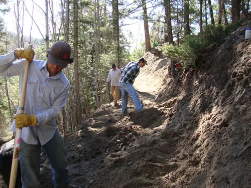 A three-person crew hand-digs a trail bench into a steep forested hillside, the nearest worker gripping a long-handled tool.