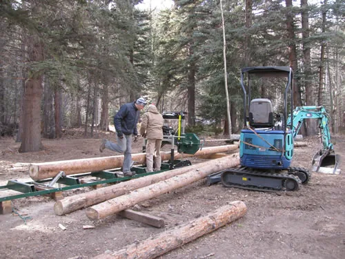 Two workers operate a portable log mill in a pine forest staging area beside a parked mini-excavator, with peeled logs stacked on the ground.