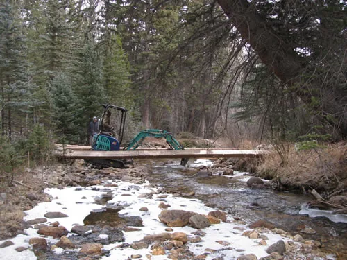 A compact tracked excavator crosses a newly set log-stringer bridge over a rocky creek, with snow patches along the banks.