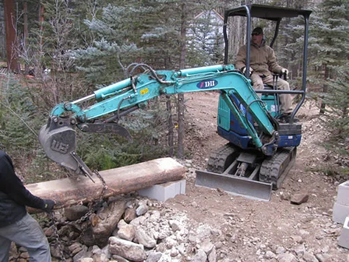 A compact tracked mini-excavator with a grapple attachment lifts a peeled log onto a freshly cut trail bench while a worker steadies the load.