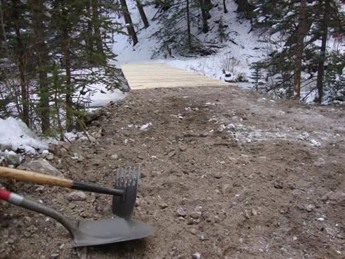 Freshly graded fine-gravel tread leads to a newly built timber bridge deck in a snowy forest, with a shovel and rake resting in the foreground.