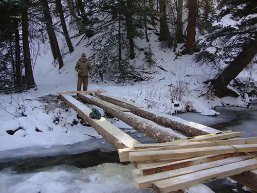 Three peeled log stringers are set across an icy creek with fresh dimensional lumber stacked in the foreground and a worker standing on the far bank.